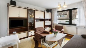 Een woonkamer met een bank, fauteuil, televisie en een raam met uitzicht op gebouwen.