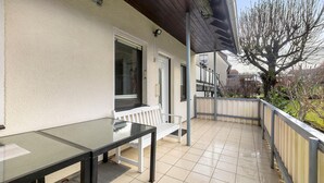 Een balkon met een glazen tafel en een witte bank, een betegelde vloer, en een boom op de achtergrond.