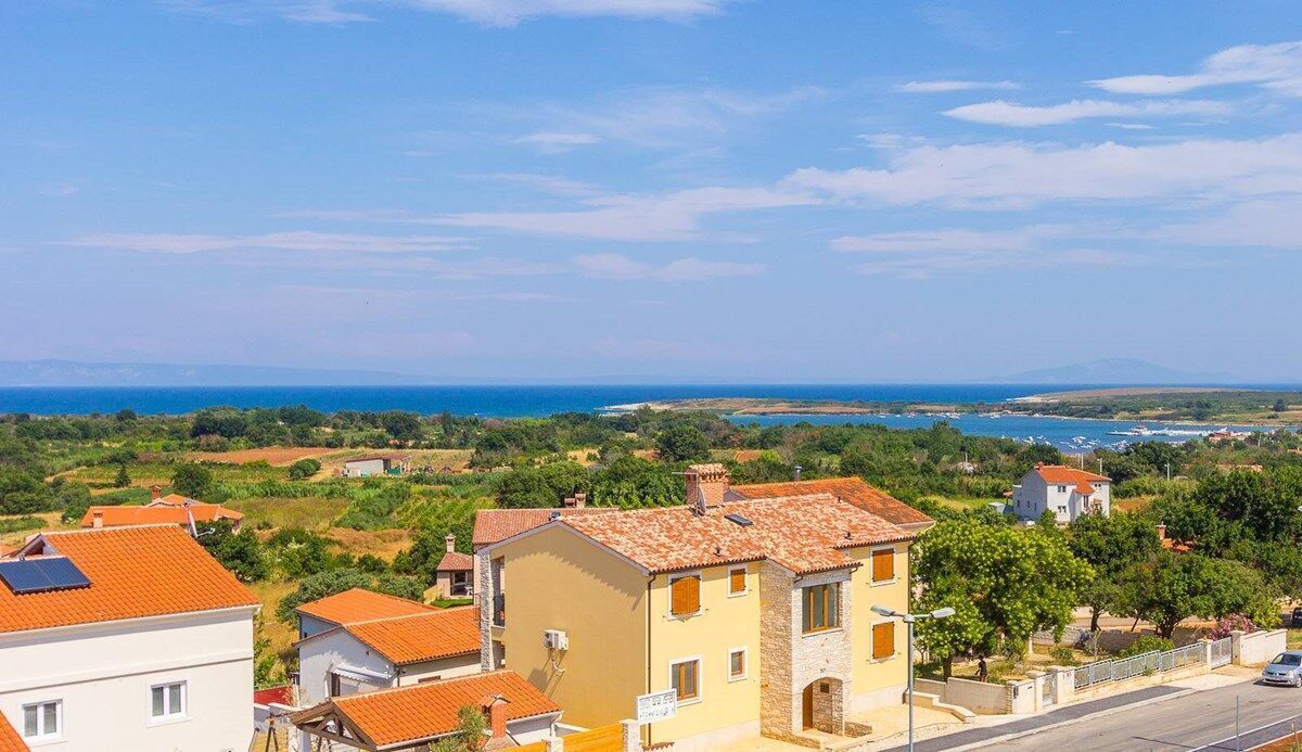 A coastal village with terracotta-roofed houses, a clear blue sky, and a view of the sea and distant mountains.