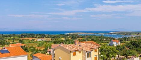 A coastal village with terracotta-roofed houses, a clear blue sky, and a view of the sea and distant mountains.