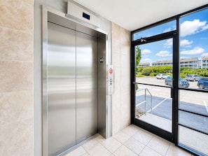 An elevator with a digital display, a parking lot view through a glass door, and a building exterior with trees and parked cars. - Smuggler's Cove 205, 2BR 2BA Oceanfront Condo in Atlantic Beach! (Atlantic Beach)