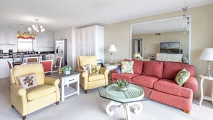 A spacious living room with a red sofa, yellow armchairs, and a glass coffee table. A kitchen with white cabinets and a dining area with a table and chairs are visible in the background. - Smuggler's Cove 205, 2BR 2BA Oceanfront Condo in Atlantic Beach! (Atlantic Beach)