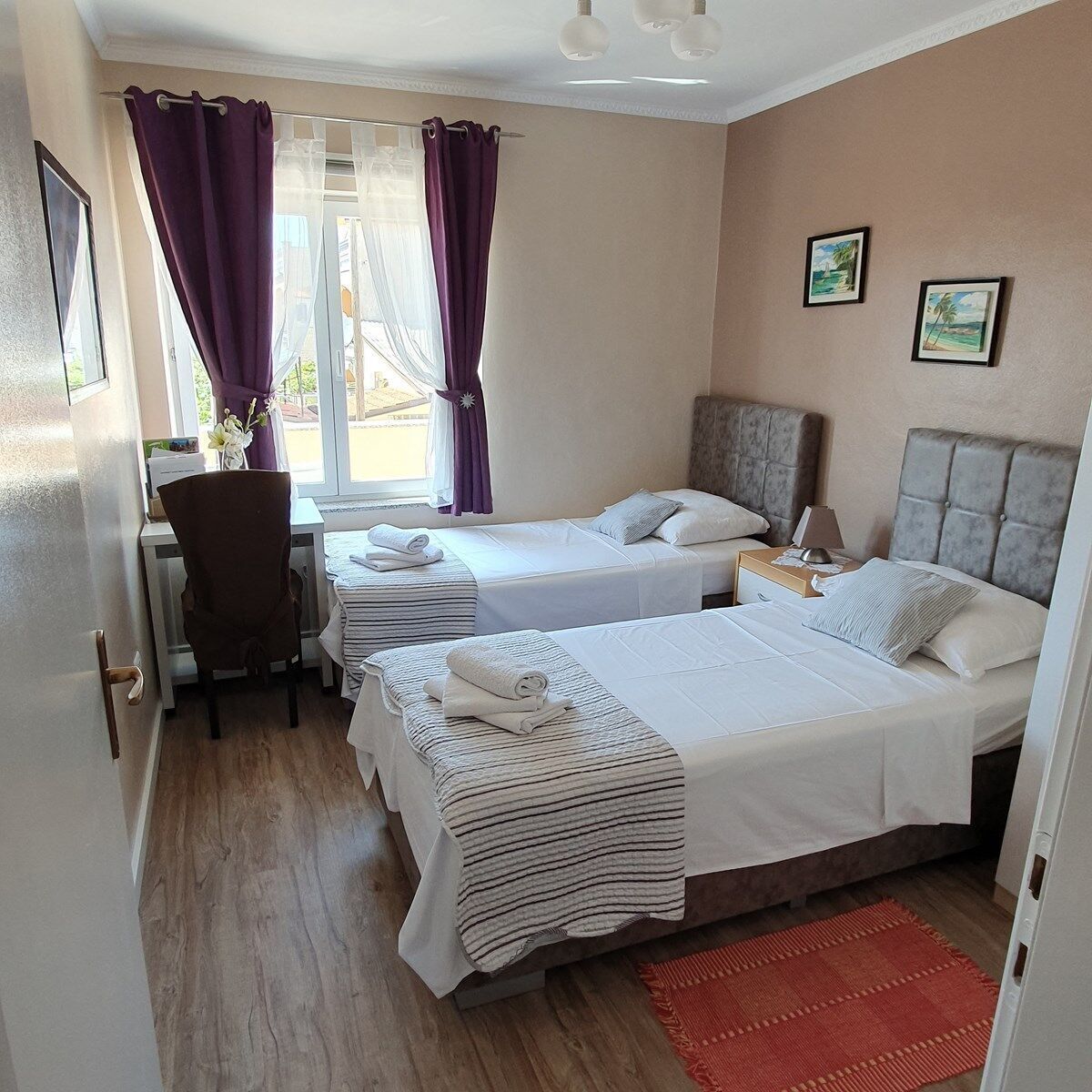 A hotel room with two beds, a desk, and a chair. There are two windows with purple curtains, a red rug, and two framed pictures on the wall.