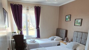 Hotelkamer met twee bedden, een bureau en een stoel. Twee ramen met paarse gordijnen, een rode vloerkleed en twee ingelijste foto’s aan de muur.