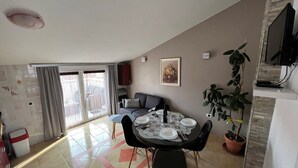 Zona pranzo con tavolo rotondo apparecchiato per quattro persone, una poltrona, una pianta in vaso e un televisore montato a parete.