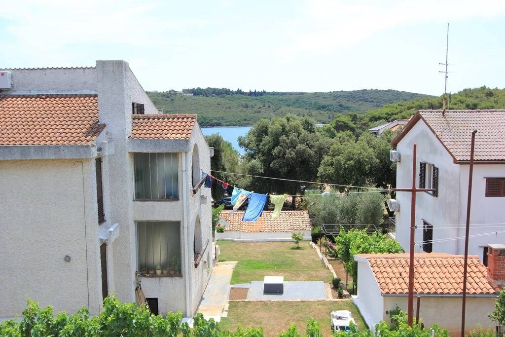 A residential area with terracotta-tiled buildings, a grassy yard, and a view of a lake and hills in the background.