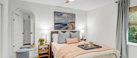 Une chambre à coucher avec un lit, un ventilateur de plafond, un tableau, une table de chevet, une lampe et une fenêtre avec des rideaux.