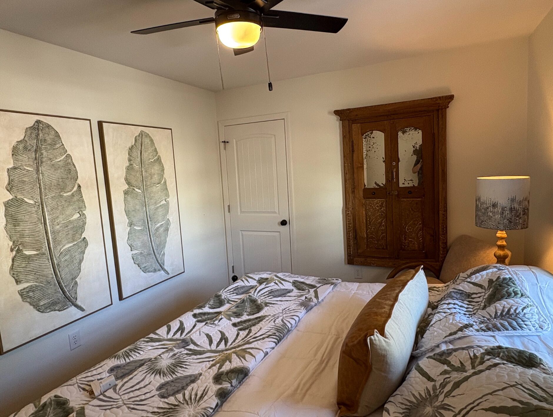 A bedroom with two single beds, a ceiling fan, a wooden cabinet, and two leaf-patterned wall art pieces.
