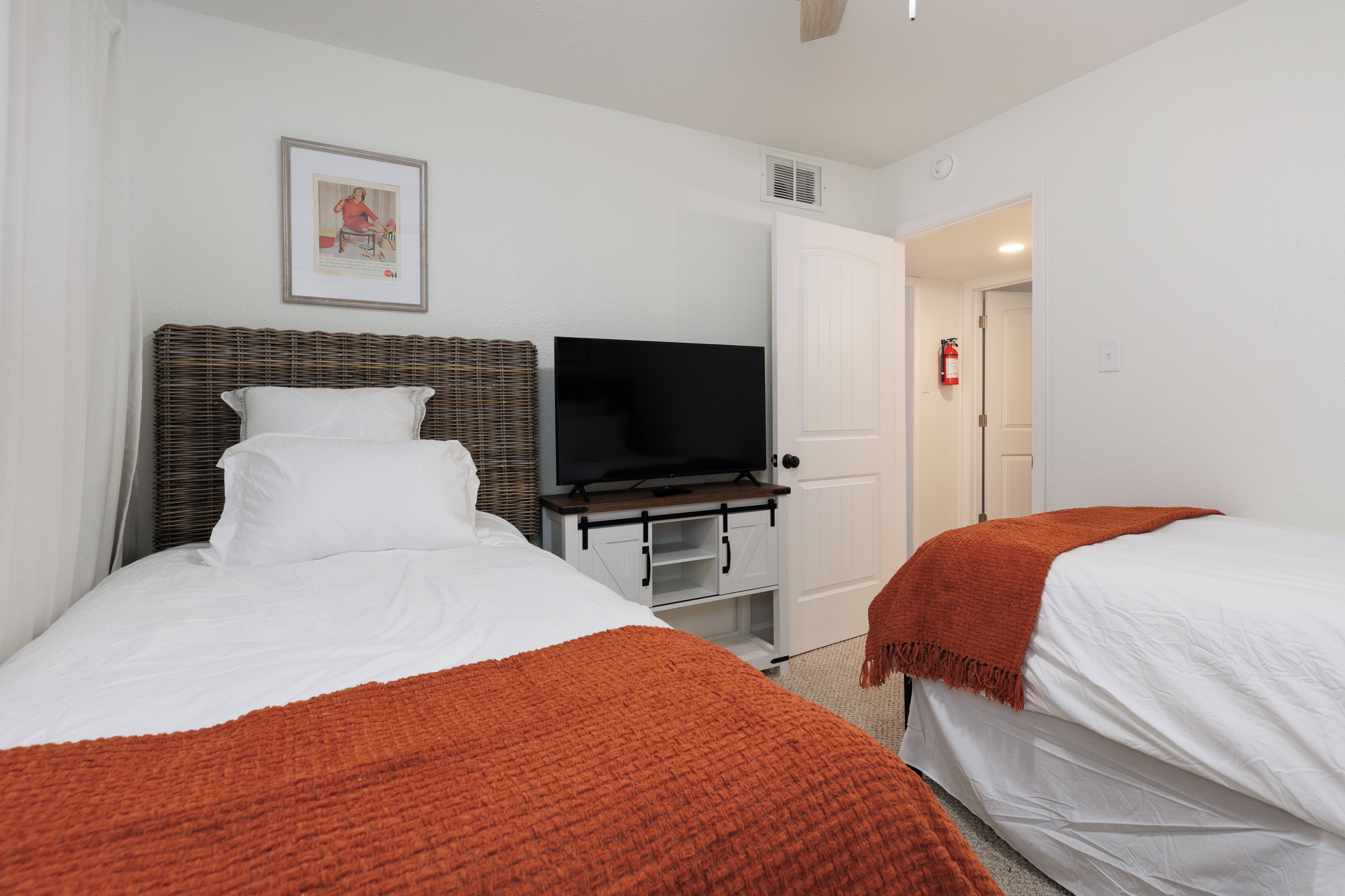 Habitación de hotel con dos camas, un televisor sobre un mueble, un cuadro en la pared y una puerta que da a otra habitación.