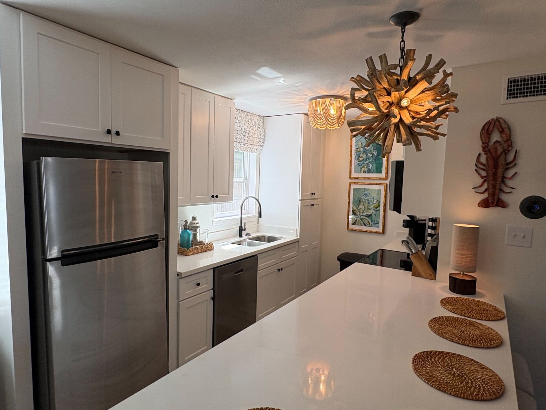 A modern kitchen with white cabinetry, a stainless steel refrigerator, a central island with woven placemats, and a unique wooden pendant light.