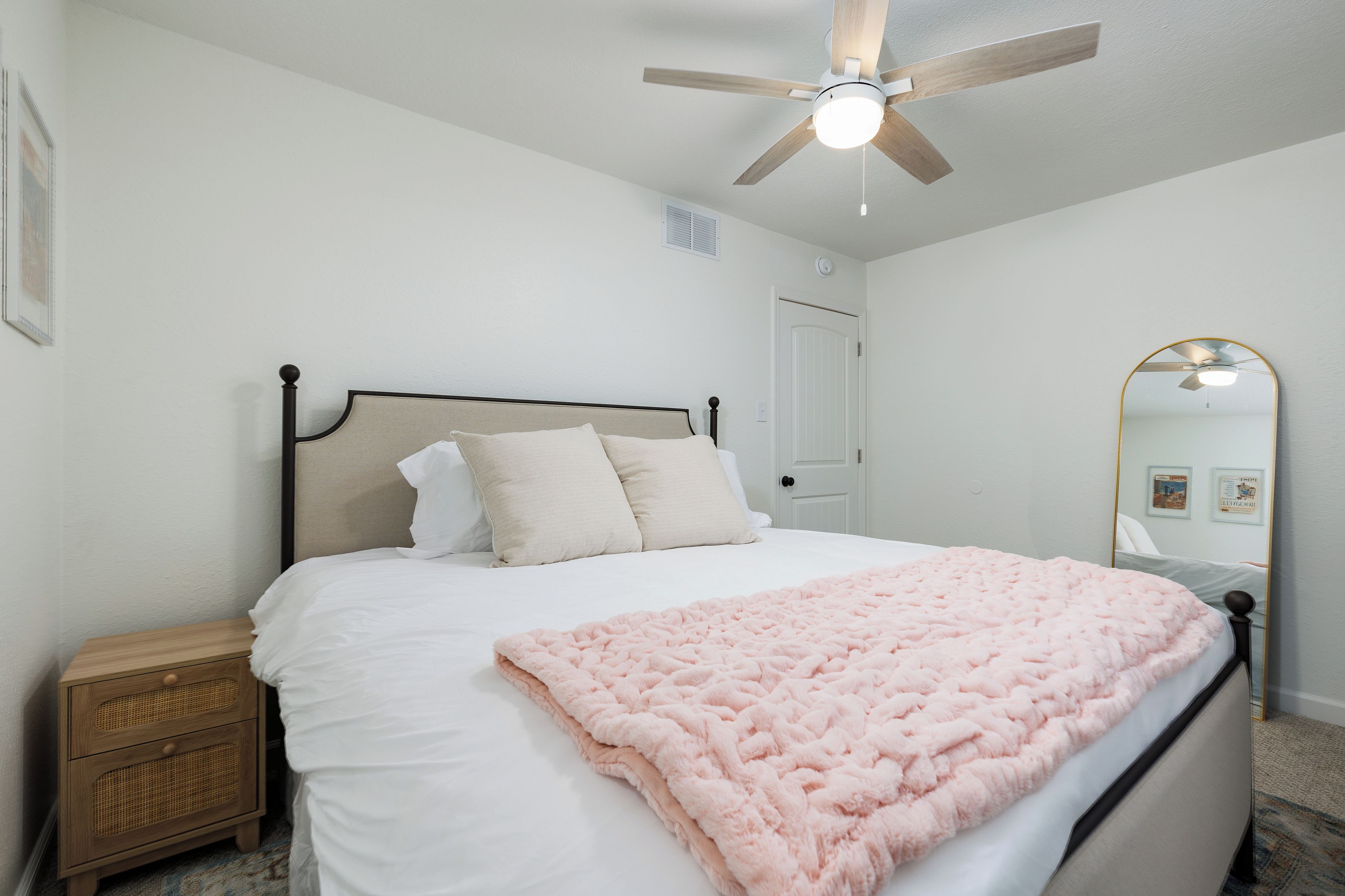 Un dormitorio con cama, mesita de noche, ventilador de techo, puerta y espejo.