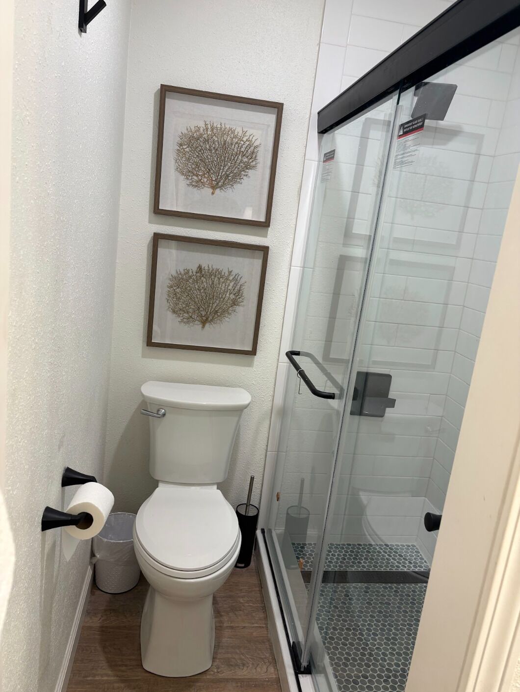 A bathroom with a toilet, two framed wall art pieces, a shower with a glass door, and a toilet paper holder.