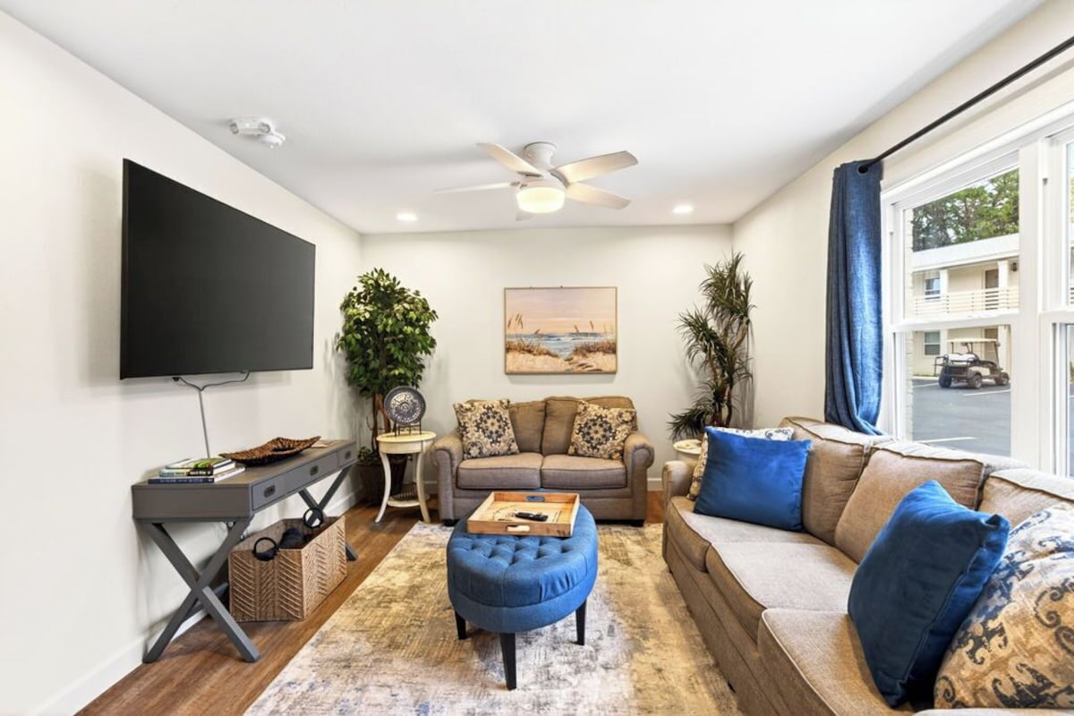 A living room with a sofa, ottoman, coffee table, TV, and artwork on the wall.