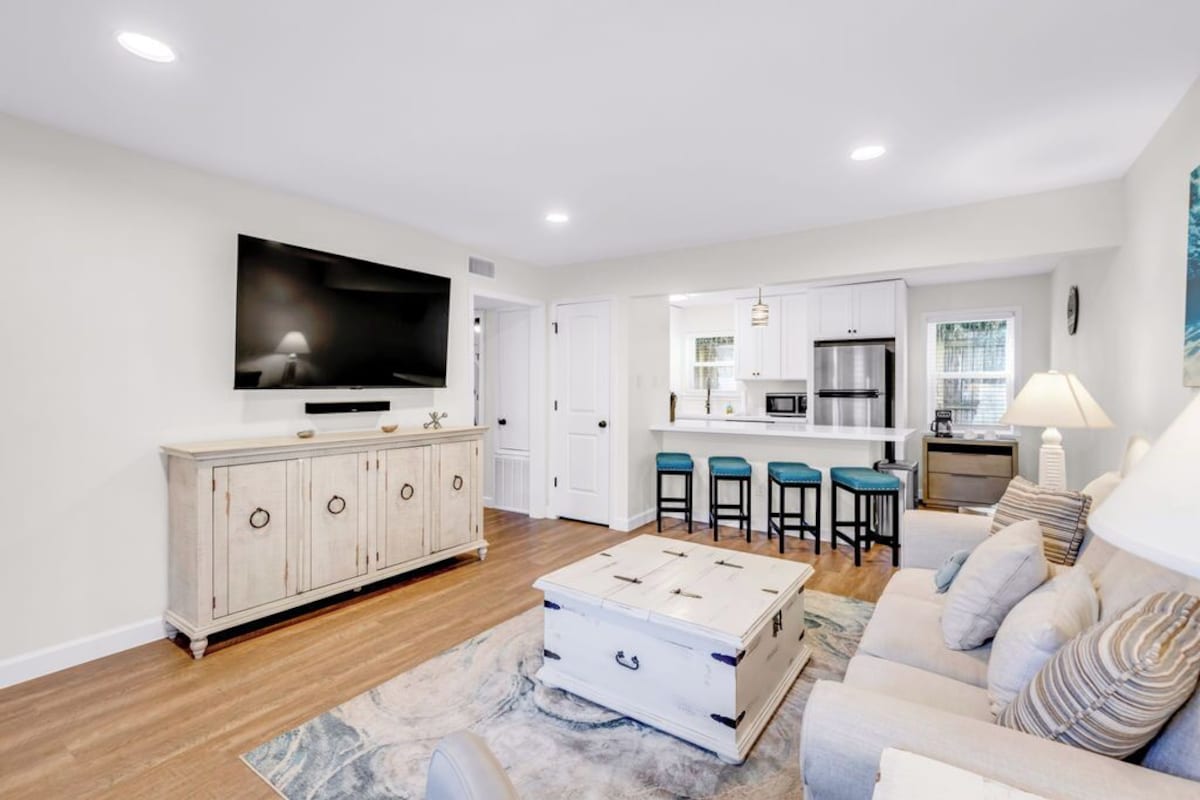 A modern living room with a flat-screen TV, a light-colored sofa, a coffee table, and a kitchen area with bar stools.