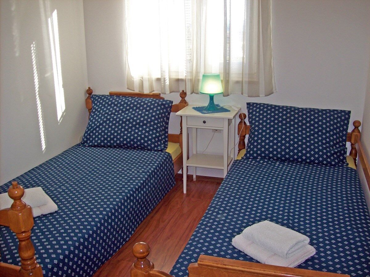 Una habitación con dos camas individuales, una mesita de noche con lámpara y una ventana con cortinas traslúcidas.
