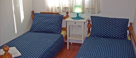 Una habitación con dos camas individuales, una mesita de noche con lámpara y una ventana con cortinas traslúcidas.