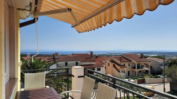 Ein Balkon mit Korbstühlen und einem Tisch, der auf ein Wohngebiet mit roten Ziegeldächern und einen ungehinderten Blick auf das Meer blickt.