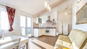 Eine moderne Küche mit weißen Schränken, einem eingebauten Backofen und einer Edelstahl-Dunstabzugshaube. Ein Essbereich mit weißem Tisch und Stühlen, und ein beiges Sofa im Wohnzimmer.