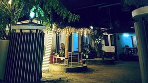 Un espace extérieur aménagé pour la nuit, comprenant un petit kiosque, un espace salon couvert et un bâtiment dont l’entrée est illuminée de lumière bleue.