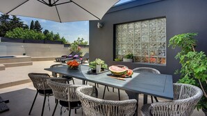 Een terras buiten met een eettafel, rieten stoelen en een grote parasol.