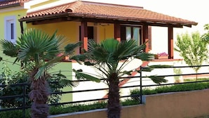 Un bâtiment à deux étages, doté d’un toit en tuiles, d’un balcon avec une balustrade et d’un petit jardin agrémenté de palmiers.
