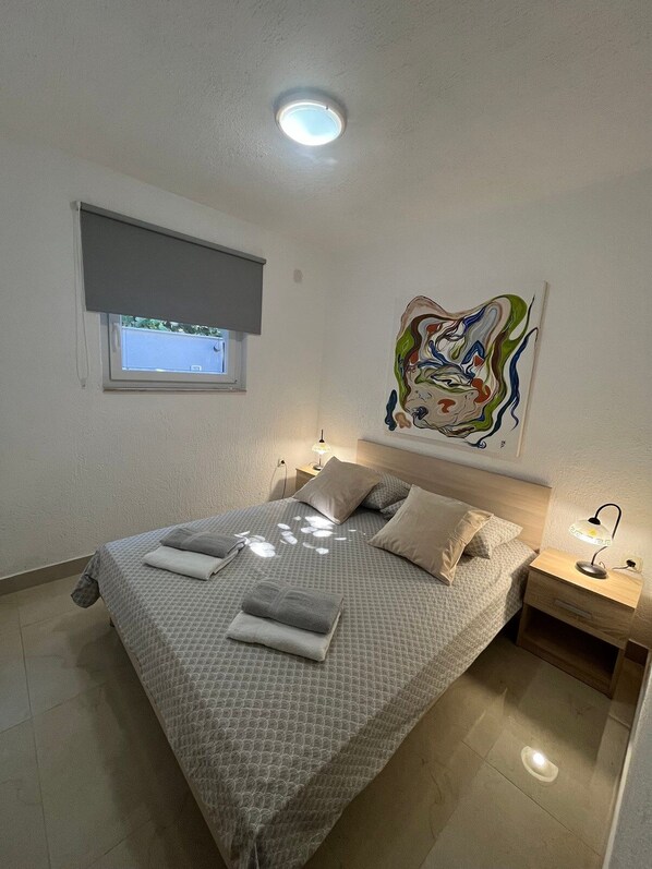 Een slaapkamer met een bed, kussens, een nachtkastje, een aan de muur hangend schilderij, een raam met een zonwering en een plafondlamp.