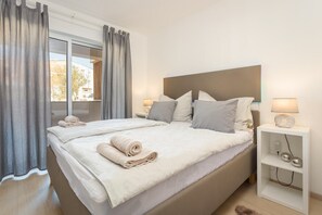 Un lit double avec une tête de lit, deux tables de chevet avec des lampes, une table de chevet blanche et une vue par une fenêtre avec des rideaux.