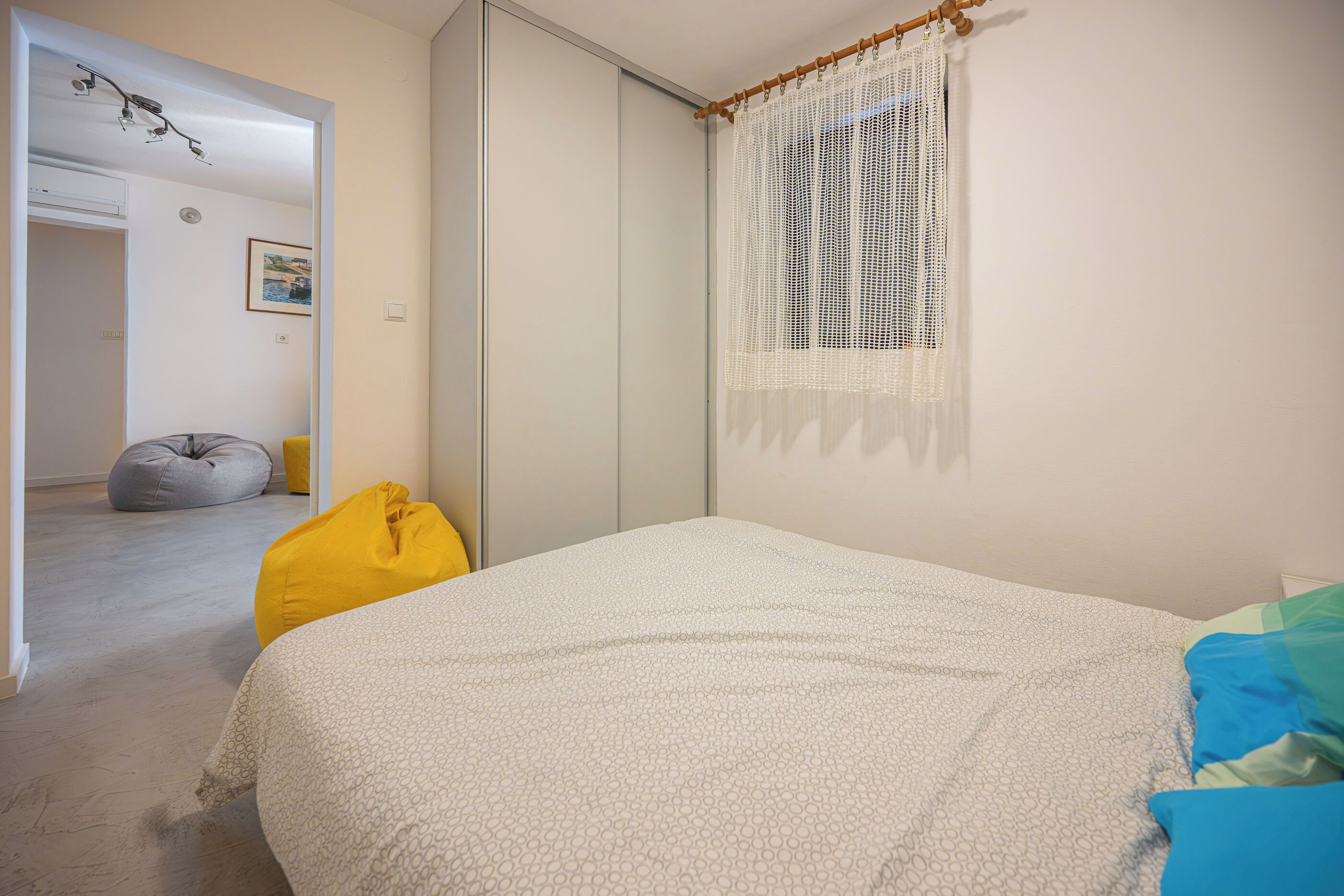 A bedroom with a bed, a wardrobe, a window with a white curtain, a bean bag, and a painting on the wall.