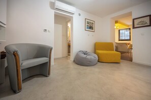 Ein Wohnzimmer mit einem grauen Sofa, einem gelben Sessel und einem grauen Sitzsack. An der Wand hängt ein gerahmtes Bild und eine Klimaanlage ist an der Wand montiert.
