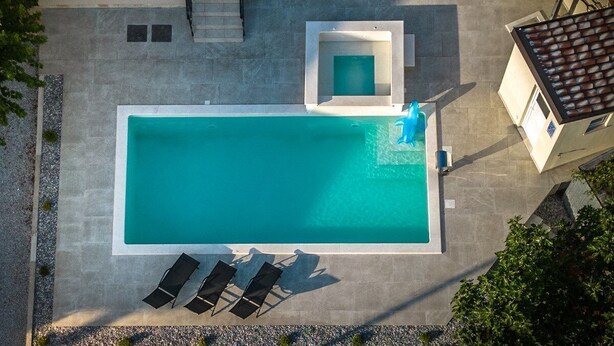 En vy från ovan av en rektangulär swimmingpool med ett litet bubbelbad, omgiven av solstolar och en trappa som leder upp till en övre nivå.
