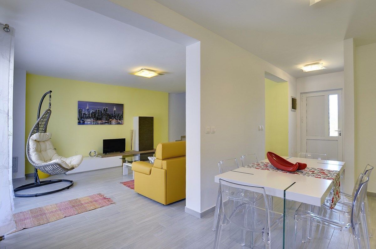 Ein modernes Wohnzimmer mit einem gelben Sofa, einem Hängekorbstuhl und einem Essbereich mit Tisch und Stühlen.