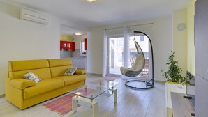 Un soggiorno moderno con un divano giallo, un tavolino in vetro, una poltrona sospesa e vista sulla cucina sullo sfondo.