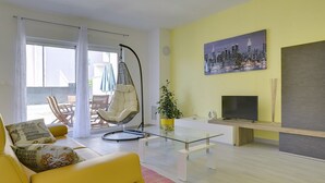 Un soggiorno con un divano giallo, una poltrona sospesa, un tavolino in vetro, una televisione e un dipinto raffigurante un panorama urbano.