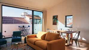 A modern living room with a sofa, chairs, a dining table, and a large window offering a view of a tiled roof and buildings. - Les Deux Mariettes Maison Rose (Lisbon)