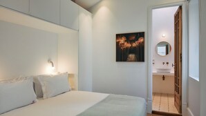 A bedroom with a bed, a wall-mounted light, a painting, and a bathroom visible through an open door. - Les Deux Mariettes Maison Rose (Lisbon)