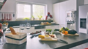 Une cuisine moderne avec des armoires blanches, un four encastré et un four à micro-ondes. On peut voir divers aliments et boissons sur le comptoir, ainsi qu’une fenêtre avec des stores et des plantes.