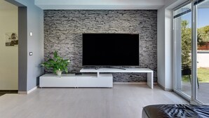 Un salon moderne avec un mur en pierre, un téléviseur à écran plat, un meuble de rangement blanc et une grande fenêtre.
