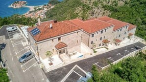 Un immeuble à plusieurs étages, doté d’un toit recouvert de tuiles rouges, d’un espace de stationnement et offrant une vue sur la mer et les collines.