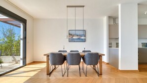 Un espace de salle à manger moderne avec une table en bois et des chaises grises, un luminaire suspendu et une vue sur l’extérieur.