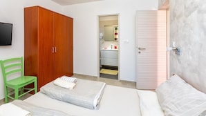 Un dormitorio con cama, una silla verde, armario de madera, baño visible a través de una puerta abierta y televisor montado en la pared.