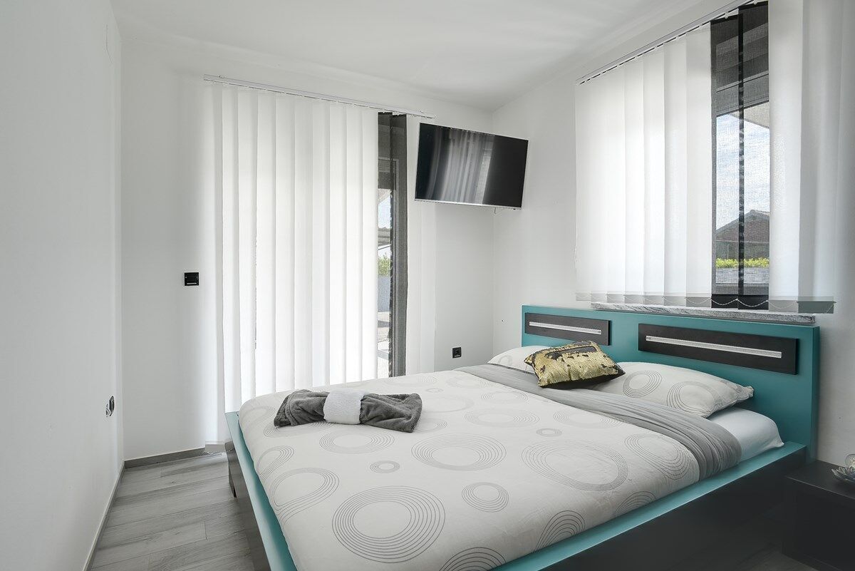 A modern bedroom with a bed, curtains, a mounted TV, and a window with blinds.