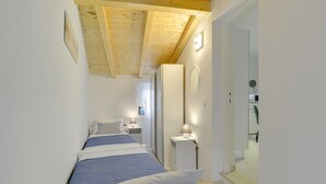 Un couloir étroit avec trois lits alignés le long du mur, une petite table de chevet avec une lampe et une porte donnant sur une salle de bain.