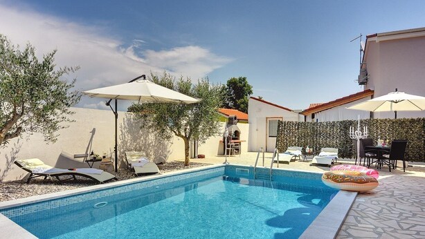 Une piscine entourée d’une terrasse en pierre et d’une haie, avec des chaises longues et des parasols.