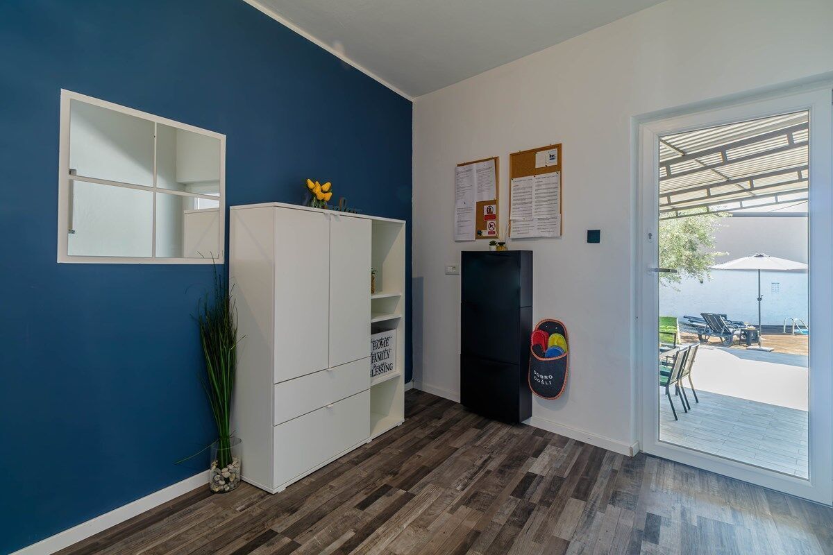 A room with a blue wall, a white cabinet, a black shelf, and a door leading to an outdoor area with a pool and lounge chairs.
