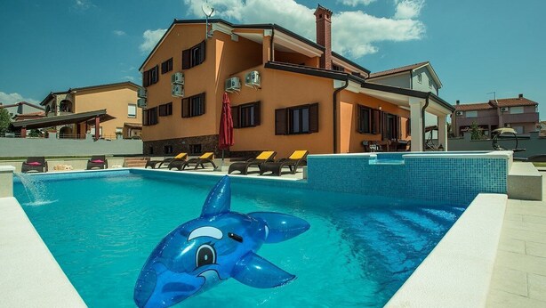 Une piscine avec un jouet gonflable en forme de dauphin, un espace détente couvert et un bâtiment doté de plusieurs balcons.