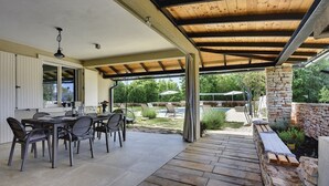 Une terrasse extérieure dotée d’un espace repas couvert, de murs en pierre et offrant une vue sur un jardin et une piscine.