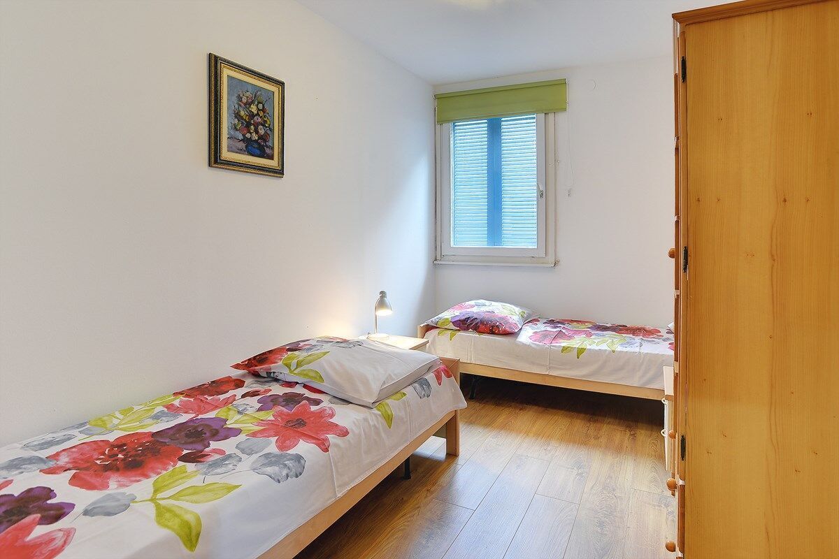 A bedroom with two single beds, a window with blinds, a wooden wardrobe, and a framed picture on the wall.