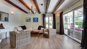 A spacious living room with a white sofa, two wicker chairs, a wooden coffee table, and a large window with curtains.