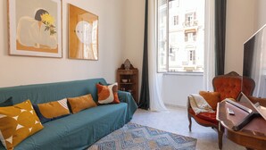 Un soggiorno con un divano color petrolio, cuscini arancioni e bianchi, un tappeto decorato e due opere d'arte incorniciate appese al muro.