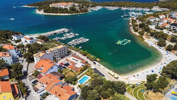 Vista aerea di una cittadina costiera con un porto turistico, spiaggia e edifici residenziali.